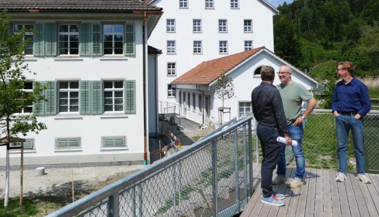 Drie mannen in gesprek op een houten terras met uitzicht op gerenoveerde witte gebouwen met groene luiken, omringd door een groene, heuvelachtige omgeving in Zwitserland